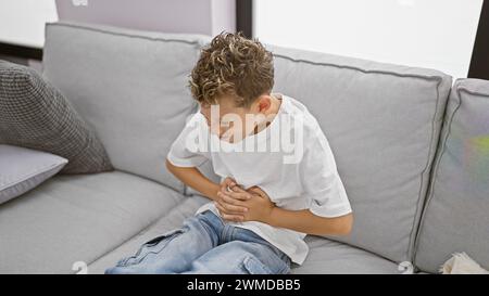 Süßer kleiner blonder Junge, der an Bauchschmerzen leidet, sitzt auf dem Sofa mit besorgtem Gesicht und berührt unruhigen Magen Stockfoto