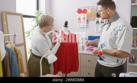 Zwei Schneider, eine Frau und ein Mann, arbeiten in einem Designraum zusammen und wählen Stoffe für ein rotes Kleid an einer Schaufensterpuppe aus. Stockfoto
