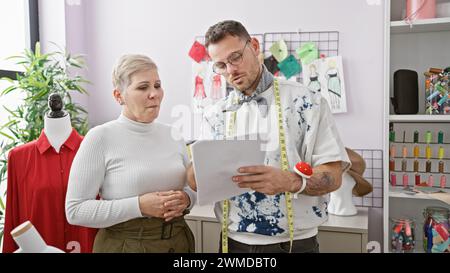Zwei Designer, ein Mann und eine Frau, arbeiten in einer modernen Schneiderwerkstatt zusammen, umgeben von Modeskizzen und Stoffrollen. Stockfoto