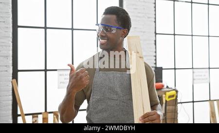 Ein lächelnder afroamerikaner, der in einer hellen Tischlerei die Daumen hochgibt Stockfoto