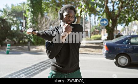 Ein junger afroamerikaner, der sich draußen auf einer Stadtstraße mit Kopfhörern und Smartwatch ausdehnt. Stockfoto