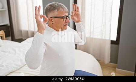 Ein Mann mittleren Alters mit grauen Haaren, Brille trägt Gesten, die drinnen auf einem Bett sitzen und eine entspannte oder verspielte Stimmung darstellen. Stockfoto