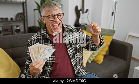 Ein aufgeregter Mann mittleren Alters, der britische Pfund hält, feiert drinnen mit einer Gitarre im Hintergrund. Stockfoto