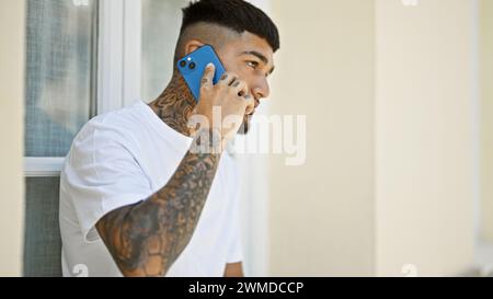 Cooler, tätowierter junger latino-Mann, der ernsthaft in Telefongespräche vertieft ist, draußen auf der sonnendurchfluteten Stadtstraße steht und entspannte Vibes ausstrahlt, während er s verwendet Stockfoto