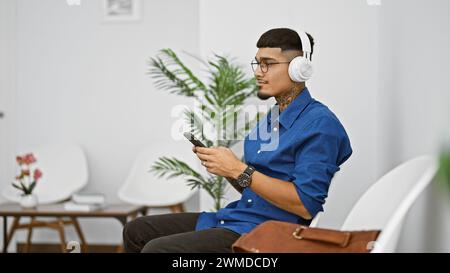 Junger lateinischer Mann, vertieft in sein Telefon, tippte dem Rhythmus, der durch seine Kopfhörer hallt, und auf einem Stuhl im Wartezimmer Stockfoto