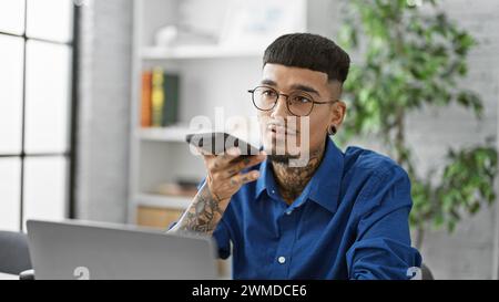 Ein junger, fokussierter lateinamerikanischer Mann, tätowierter Geschäftsarbeiter, der in einem erfolgreichen Büroporträt festgehalten wurde und Sprachnachrichten per Smartphone an seinem Arbeitsplatz versendet Stockfoto