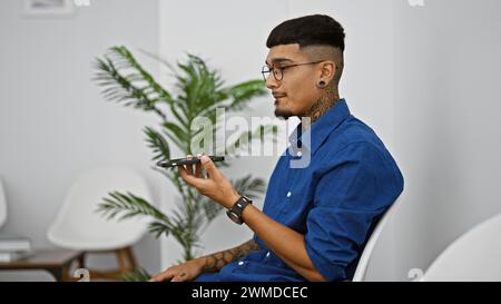 Junger lateinamerikanischer Mann, der in ernsthafte Gespräche vertieft ist, eine Sprachnachricht über sein Smartphone sendet und bequem im Wartezimmerstuhl sitzt Stockfoto