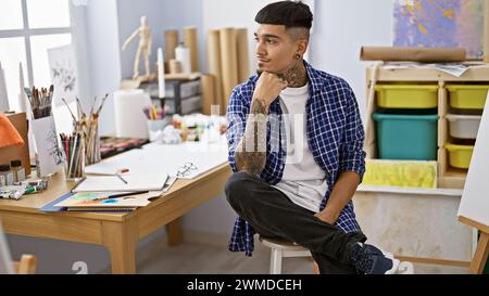 Tätowierter junger lateinamerikanischer Mann, ein vertiefter Künstler, der mit ernstem Gesicht in einem Kunststudio an seiner Staffelei sitzt, mit Pinsel in der Hand Stockfoto