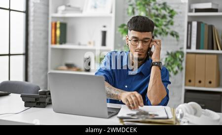 Hübscher junger lateinmann, ein seriöser Geschäftsarbeiter, der in die Online-Arbeit auf seinem Laptop vertieft ist und sich in ein hitziges Telefongespräch mit seinem Smart konzentriert Stockfoto