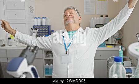 Mann mittleren Alters im Laborkitt, der sich in einem Labor dehnt, zufrieden ist und eine Arbeitsunterbrechung zeigt. Stockfoto