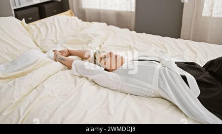 Hispanische Frau mit kurzen blonden Haaren, die auf dem Rücken auf einem Bett in einem gut beleuchteten Schlafzimmer liegt und entspannt und besinnlich aussieht Stockfoto