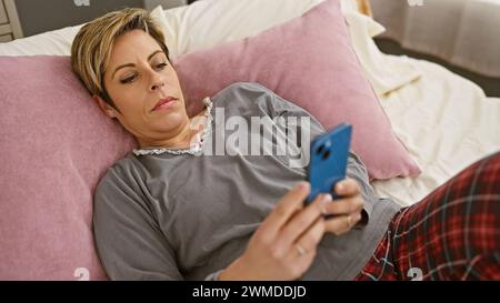 Hispanische Frau mit kurzen blonden Haaren, die ein Smartphone im Schlafzimmer benutzt und an Freizeit- und Technikthemen erinnert. Stockfoto