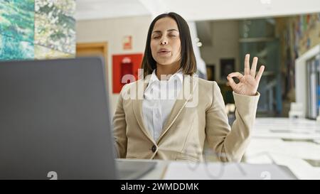 Eine junge hispanische Frau meditiert, während sie einen Laptop in einem isolierten gelben Hintergrund benutzt. Stockfoto