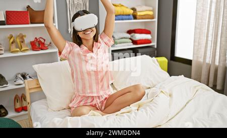 Eine fröhliche hispanische Frau im Pyjama verwendet ein Virtual-Reality-Headset in einem modernen Schlafzimmer und drückt Begeisterung und Technologie aus. Stockfoto