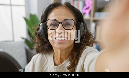 Hispanische Frau in ihren 50ern, macht ein Selfie drinnen mit Kopfhörern, lächelt in die Kamera. Stockfoto