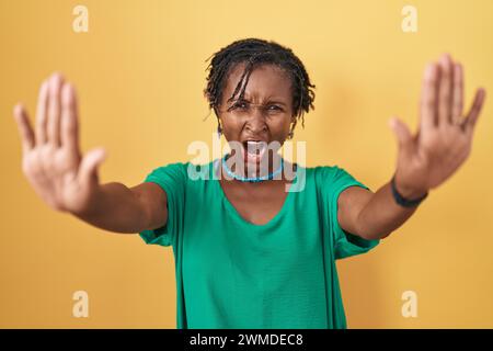 Afrikanische Frau mit Dreadlocks, die über gelbem Hintergrund stehen, stoppt mit Handflächen, wütend und frustrierend Stockfoto