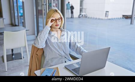 Eine nachdenkliche junge Frau mit Brille arbeitet an einem Laptop in einem Café im Freien in der Stadt. Stockfoto