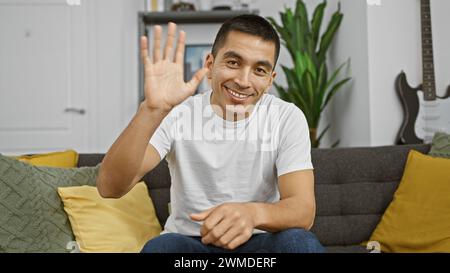 Ein lächelnder junger hispanischer Mann in lässiger Kleidung winkt Hallo, während er drinnen auf einer Couch sitzt. Stockfoto
