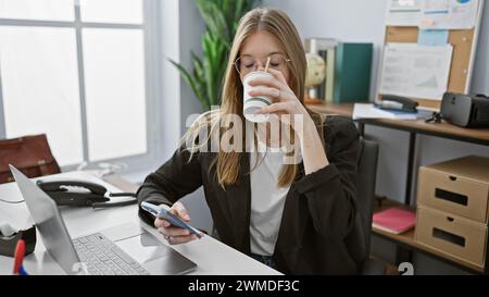 Blonde Geschäftsfrau trinkt Kaffee und benutzt Smartphone in moderner Büroumgebung Stockfoto