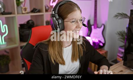 Lächelnde weiße Frau mit Kopfhörern in einem neonbeleuchteten Spielzimmer in der Nacht, die glücklich mit ihrem Computer interagiert Stockfoto