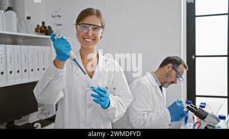 Zwei fröhliche Laborkollegen, Frau und Mann, gießen zusammen Flüssigkeit in eine Probenpipette, navigieren auf dem Weg der medizinischen Wissenschaft, Handschuhe für Sicherheit in A Stockfoto