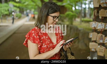 Wunderschöne hispanische Frau in Brille, die Gebetstafeln im japanischen Gotokuji-Tempel festnimmt und die Tradition durch ein Smartphone-Objektiv bewahrt Stockfoto