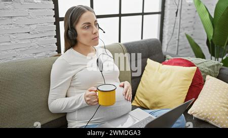 Schwangere hispanische Frau im Kopfhörer sitzt mit einem Laptop in einem gemütlichen Wohnzimmer und hält einen gelben Becher Stockfoto