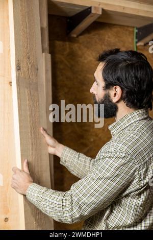 Vertikal fotokonzentrierter Zimmermann sortiert Holzdielen und wählt das perfekte Stück für sein nächstes Projekt in einem gut organisierten Holzlager aus Stockfoto