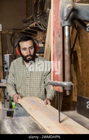 Vertikales Foto ein aufmerksamer Zimmermann mit Gehörschutz führt vorsichtig ein Holzbrett durch eine Bandsäge und fokussiert dabei intensiv Stockfoto