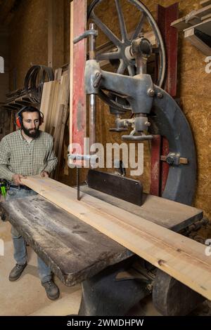 Vertikales Foto ein Meister des Zimmermannes, mittelerwachsener kaukasier, mit konzentrierter Entschlossenheit, speist eine lange Holzplatte durch eine alte Bandsäge, umrahmt Stockfoto