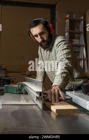 Vertikales Foto ein konzentrierter Zimmermann A mit Gehörschutz verwendet eine Tischsäge, um Holz zu schneiden, was sein Know-how und seine Liebe zum Detail zum Ausdruck bringt.Copy Spac Stockfoto