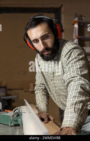 Ein in seine Arbeit vertieftes vertikales Foto führt ein Zimmermann mitten im Erwachsenenalter kaukasier mit Sicherheits-Ohrenschützern akribisch ein Stück Holz durch einen Tisch sa Stockfoto