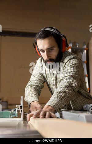 Ein aus einem dynamischen Winkel aufgenommenes vertikales Foto wird von einem Zimmermann mittlerer Erwachsener mit Gehörschutz vollständig absorbiert, wenn eine Tischsäge bedient wird Stockfoto