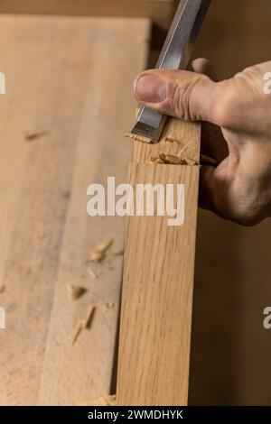 Vertikales Foto das Foto konzentriert sich auf die präzise Handbewegung eines Zimmermanns, während eine Kerbe in eine Holzdiele gemeißelt wird, wobei Holzspäne verstreut sind. C Stockfoto