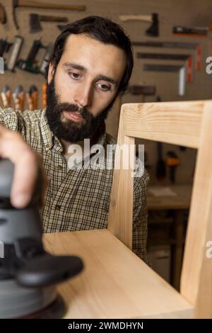 Vertikaler fotokonzentrierter Zimmermann mittlerer Erwachsener kaukasischer, der einen Holzstuhl mit einem tragbaren Schleifgerät in einem Well-e zu einem glatten Finish abschleift Stockfoto