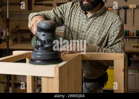 Ein horizontaler fotofokussierter Holzarbeiter in einem karierten Hemd verwendet einen Schwingschleifer auf einem Stuhl, der in einem gut ausgestatteten Studio ein glattes Finish schafft. Busine Stockfoto