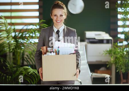 Neuer Job. Lächelnde, moderne 40-jährige Arbeiterin im modernen grünen Büro im grauen Business-Anzug mit persönlichen Sachen im Karton. Stockfoto