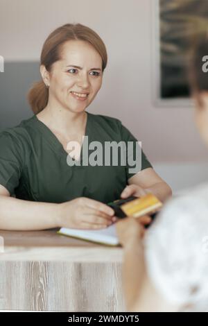 Gesundheitszeit. Lächelnde Massage-Therapeutin im Massageschrank, der Kunde akzeptiert Kreditkartenzahlungen über das Terminal an der Rezeption. Stockfoto