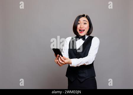 Aufgeregt glückliche asiatische Hotelrezeptionistin in Uniform mit Smartphone und Blick in die Kamera. Überraschte, lächelnde junge Kellnerin, die gute Nachrichten auf dem Handy-Porträt liest Stockfoto