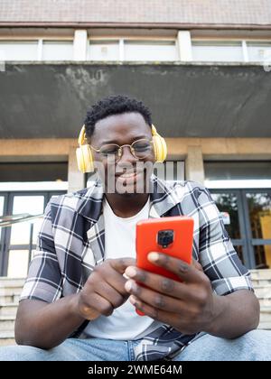 Junger afroamerikanischer Mann mit Brille und Kopfhörern, der mit Handy auf dem Campus plaudert Stockfoto