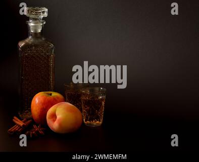 Apfelpfirsichlikör mit Zimt und Anis auf schwarzem Hintergrund, hausgemachter Alkohol in einer Karaffe und Gläser mit Gewürzen und Reifen Früchten. Nahaufnahme. Stockfoto