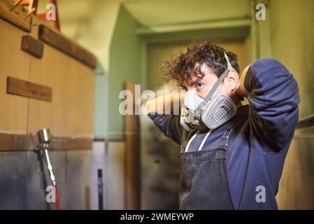 Porträt eines jungen hispanischen Arbeiters, der eine Atemschutzmaske entfernt, während er seine Spritzlackierarbeiten in seiner Werkstatt beendet. Echte Menschen bei der Arbeit. Stockfoto