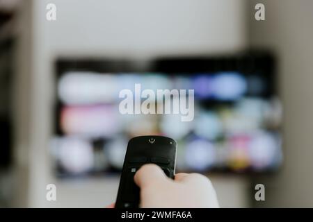 Hand hält die Fernbedienung, die auf den Fernseher zeigt. Person, die Smart-TV in Innenräumen ansieht. Unscharfer Hintergrund. Farbenfroher Bildschirm. Verwalten Sie Geräte, Elemente eines Smart Home Control Panels, Komfort fürs Leben Stockfoto