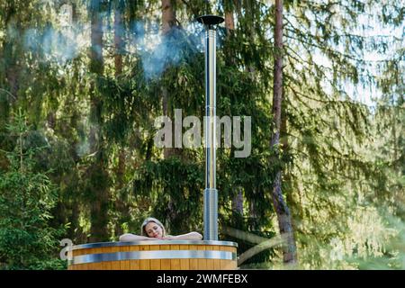 Junge Frau, die in der Holzbadewanne mit Kamin Holz verbrennt und Wasser im Hinterhof in den Bergen erwärmt. Stockfoto