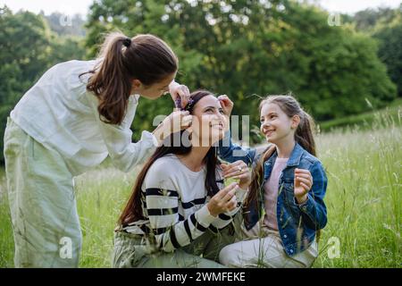 Schöne Mutter mit Töchtern, Blumen pflücken, Haare einziehen, im Gras auf der Wiese sitzen. Muttertag und mütterliche Liebe. Stockfoto