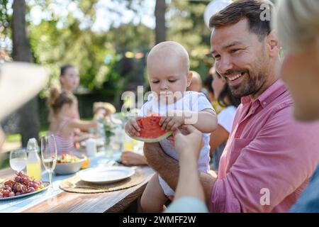Süßes Baby isst Wassermelone auf einer Gartenparty im Sommer. Dad hält den Jungen in den Armen und lächelt. Stockfoto