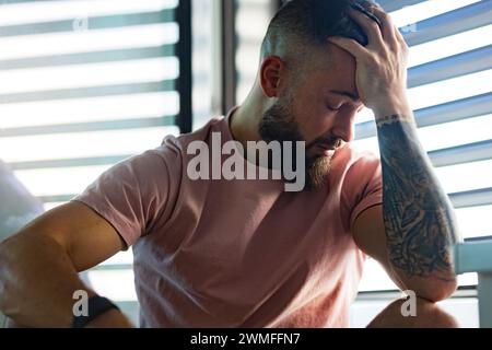 Ängstlicher Mann in rosa T-Shirt mit Tätowierung auf den Armen, der Kopf in der Hand hält. Stockfoto