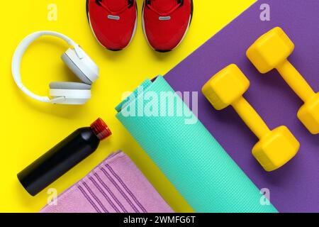 Fitnessgeräte. Sporttrainingskonzept. Kurzhanteln auf lila Rollenmatte auf gelbem Hintergrund. Gesunder Lebensstil. Flache Lagen. Rote Sneakers, Musik-Headpho Stockfoto