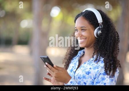 Glückliche schwarze Frau, die Kopfhörer trägt und Musik hört und draußen ihr Telefon hält Stockfoto