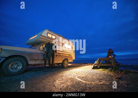 Frau, die auf einem Picknicktisch neben einem Wohnmobil sitzt, auf einem Surftrip Stockfoto